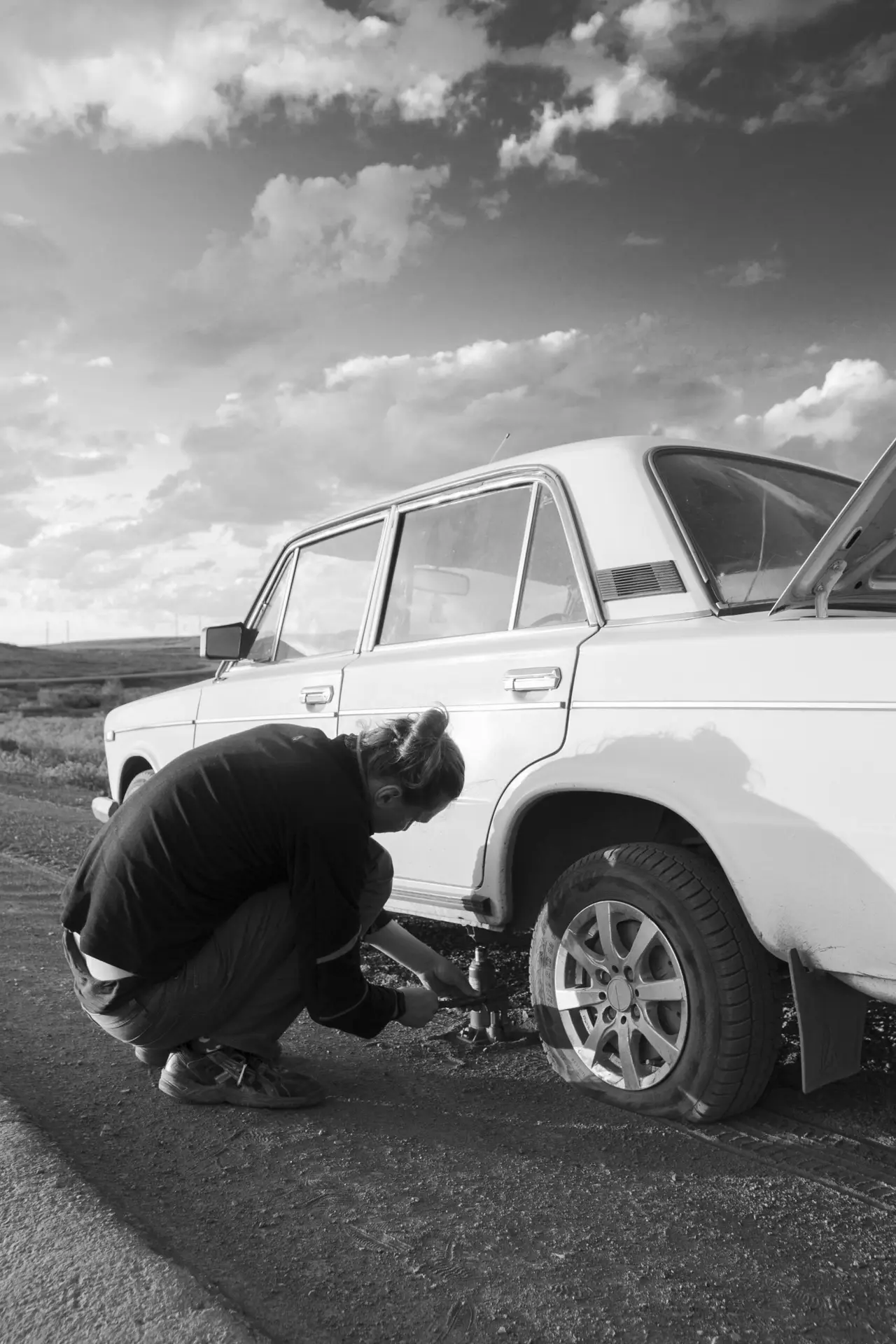 A man changes a car tire on a country road. Black and white photo.
