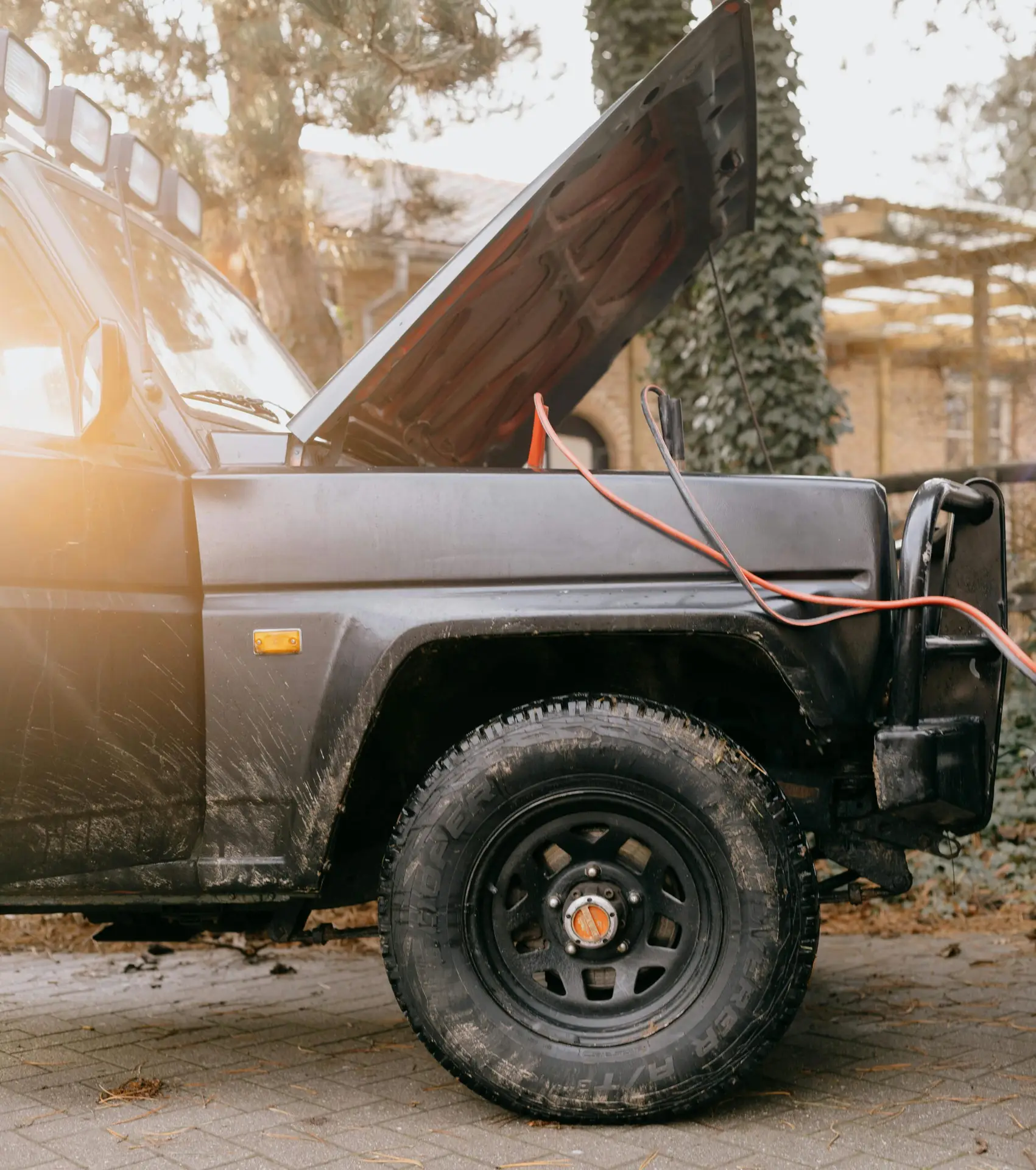 Close-up of black SUV with hood open and jumper cables, set outdoors on a sunny day.